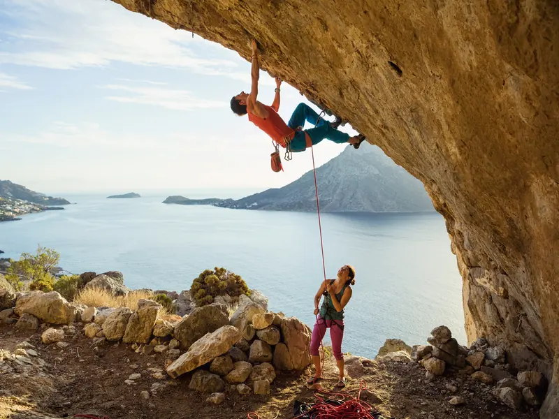 A climber hanging from a sunlit cave overhang above the sea while his partner belays and watches from below