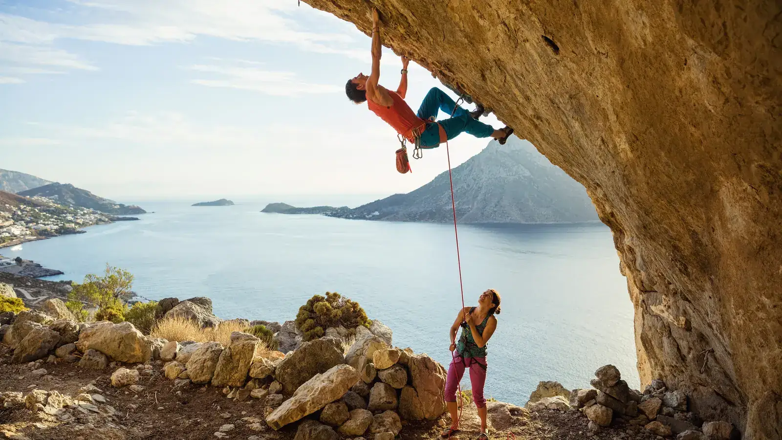 A climber hanging from a sunlit cave overhang above the sea while his partner belays and watches from below