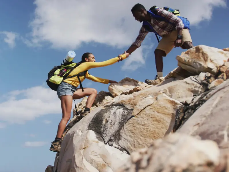 Two hikers with backpacks scrambling up a sunny granite boulder together