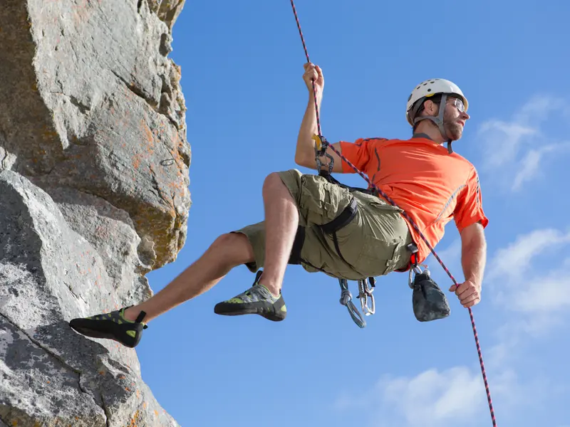 A smiling climber in a helmet rappelling down a rock face against a bright blue sky