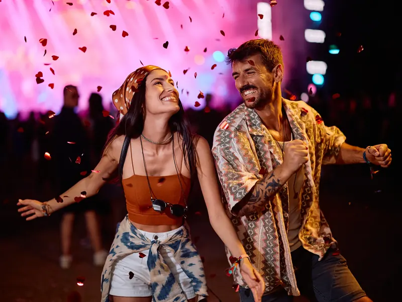 A laughing couple dancing together at a music festival under falling heart confetti