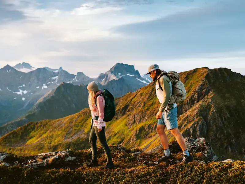 Two hikers with backpacks walking along a grassy mountain ridge with snow-capped peaks behind them