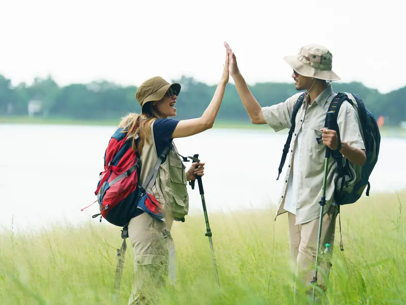 Two hikers with trekking poles and backpacks high-fiving in a meadow beside a lake