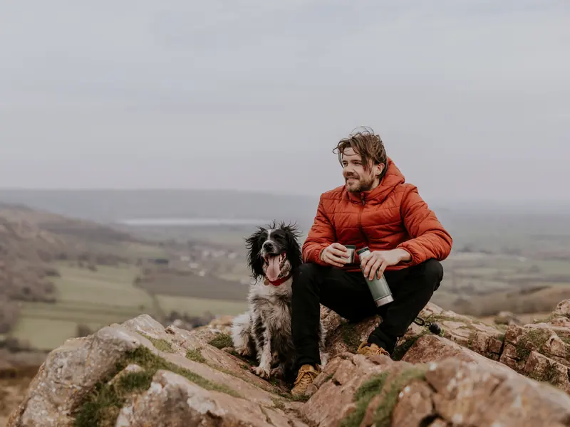 A man in a red jacket resting on a rocky hilltop beside his dog