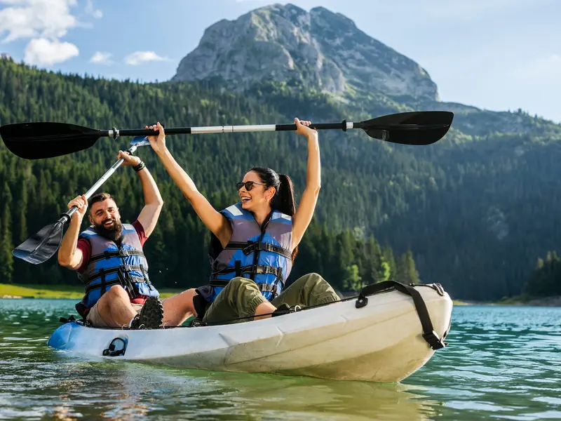 A couple in lifejackets sitting in a tandem kayak on a mountain lake, both raising their paddles and smiling