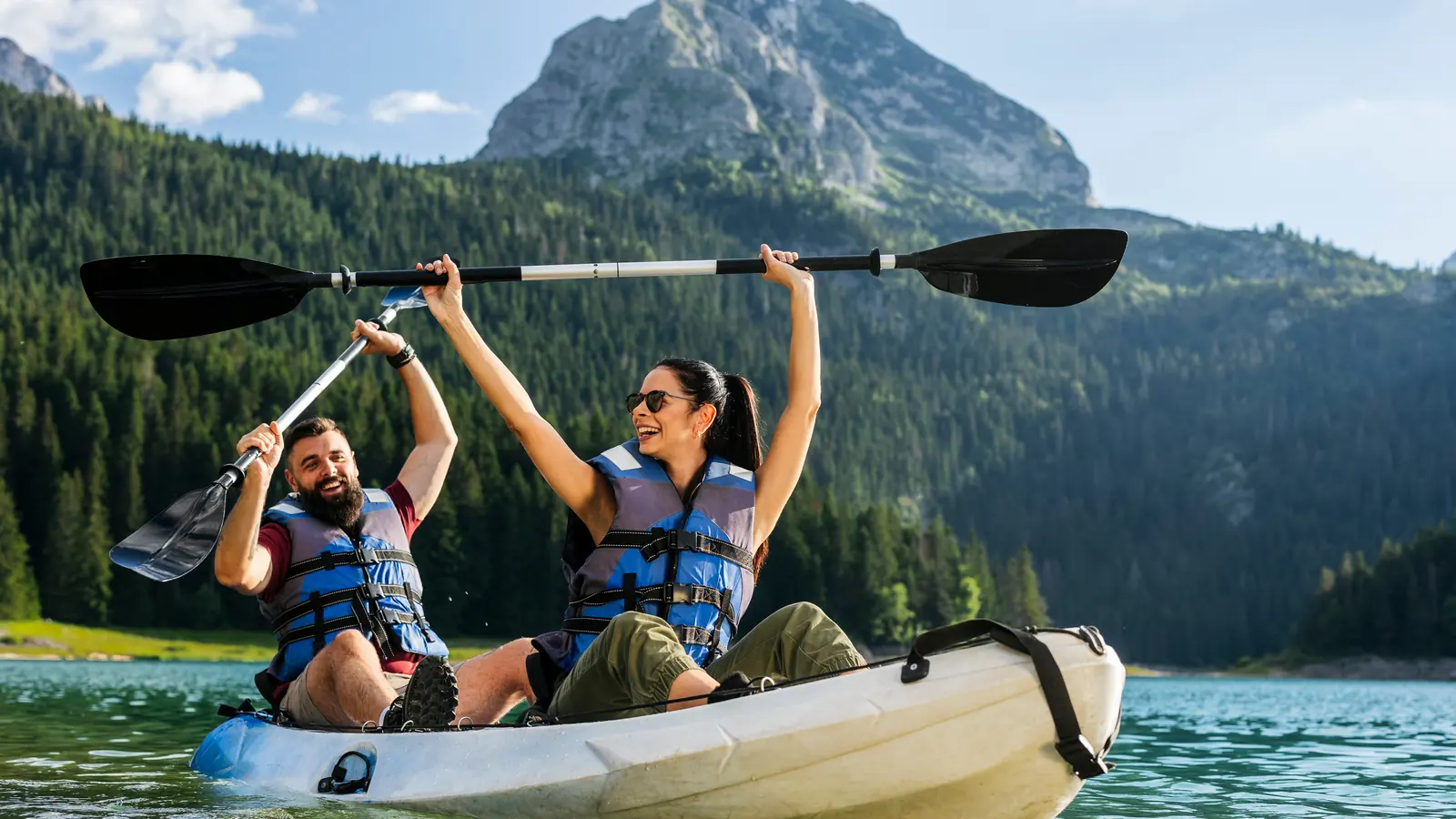 A couple in lifejackets sitting in a tandem kayak on a mountain lake, both raising their paddles and smiling