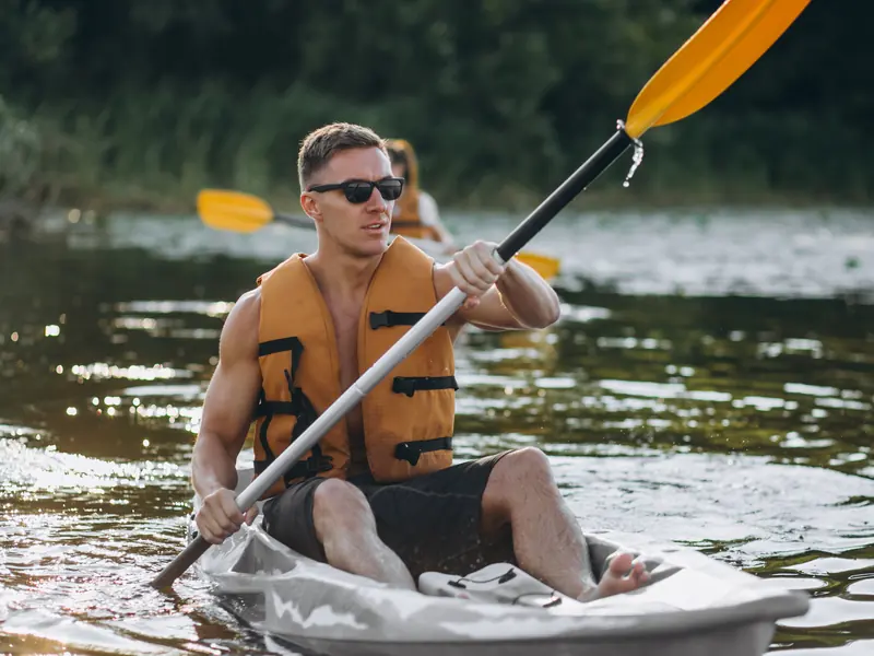 A man in sunglasses and an orange lifejacket paddling a kayak on a sunlit river