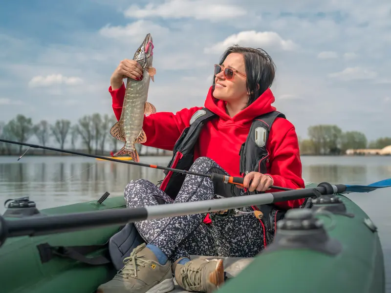 A smiling woman in a red hoodie holding up a pike she caught from her inflatable kayak