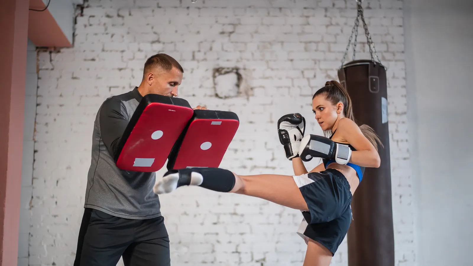 A woman in boxing gloves landing a high kick on pads held by her training partner in a brick-walled gym