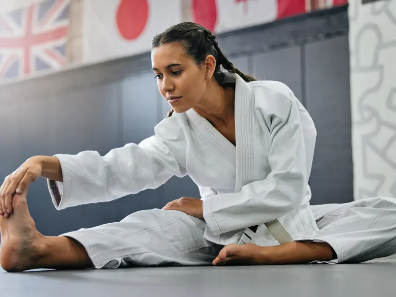 A woman in a white gi stretching toward her foot on a dojo mat