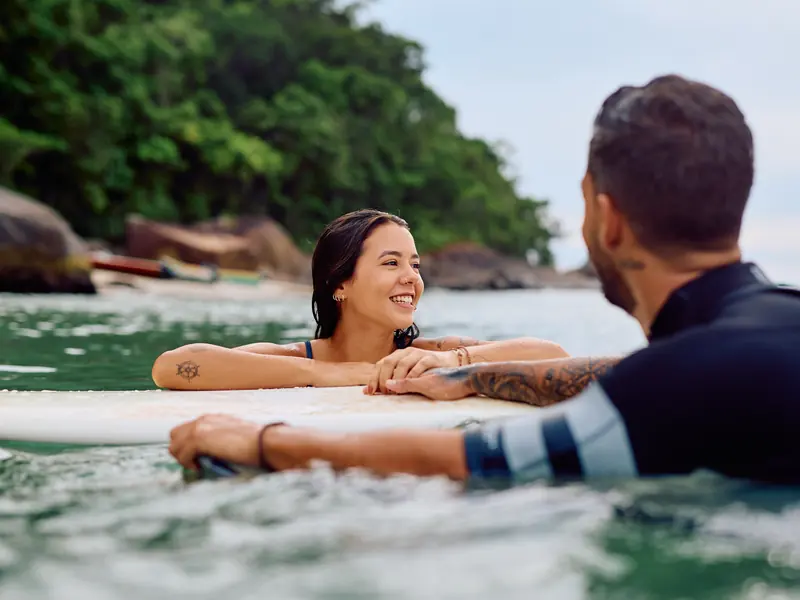 A smiling woman resting her arms on a board in the sea while a man in a wetsuit floats beside her