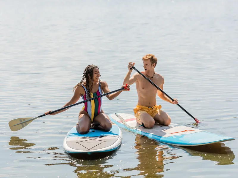 A laughing couple kneeling side by side on paddleboards on a calm lake
