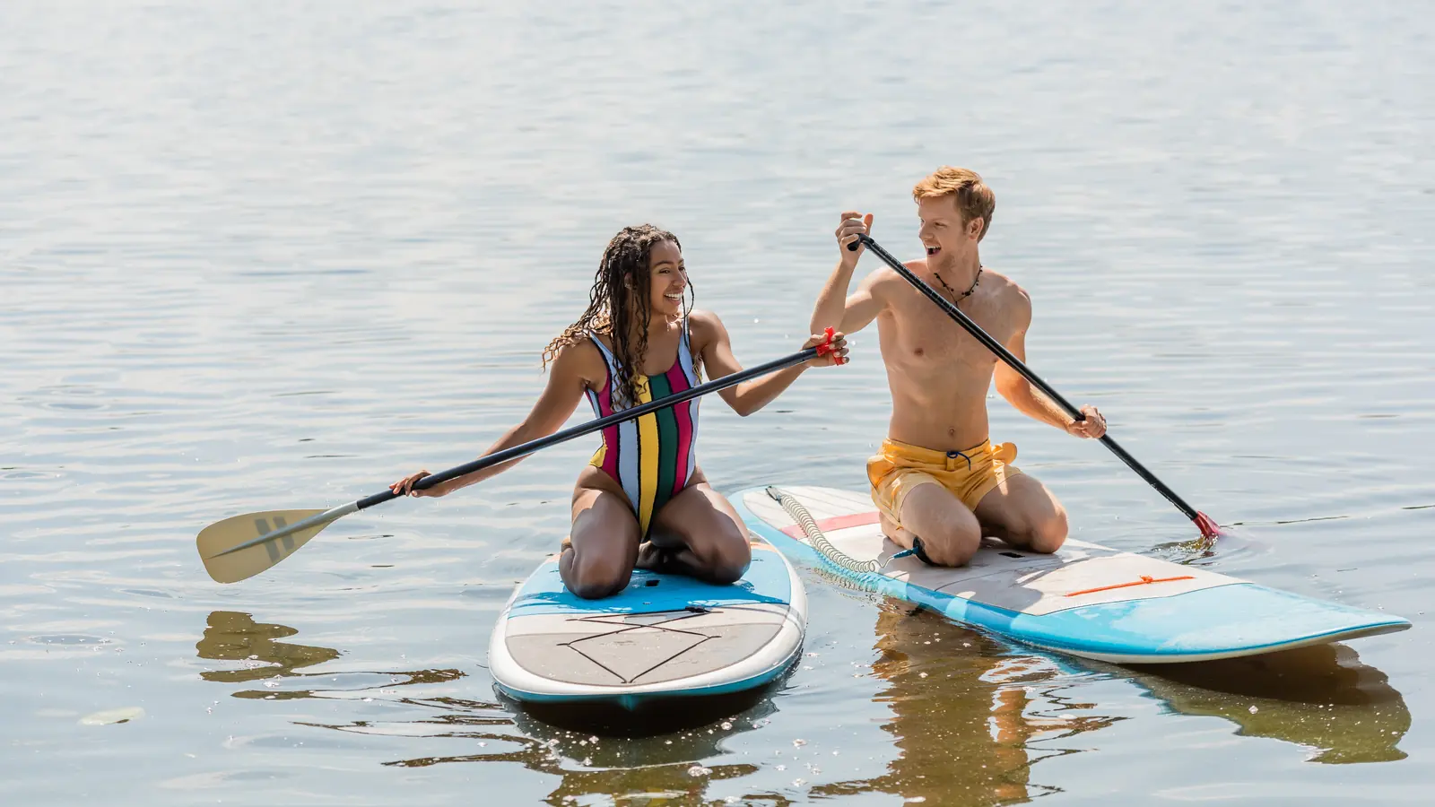 A laughing couple kneeling side by side on paddleboards on a calm, sunlit lake