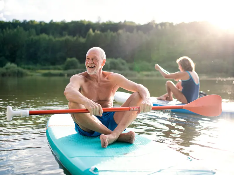 A laughing silver-haired man sitting on a paddleboard on a sunny lake while a woman paddles behind him