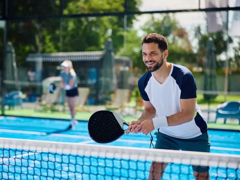 A smiling man with a padel racket ready at the net on a sunny blue court