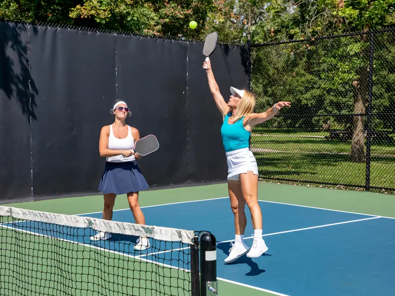 Two women playing doubles on a sunny outdoor court, one leaping to smash the ball