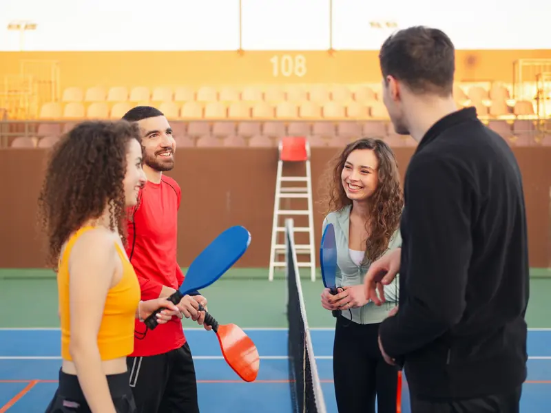A group of friends holding pickleball paddles, laughing and chatting at the net