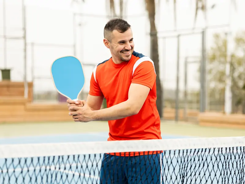 A smiling man holding a blue pickleball paddle at the net