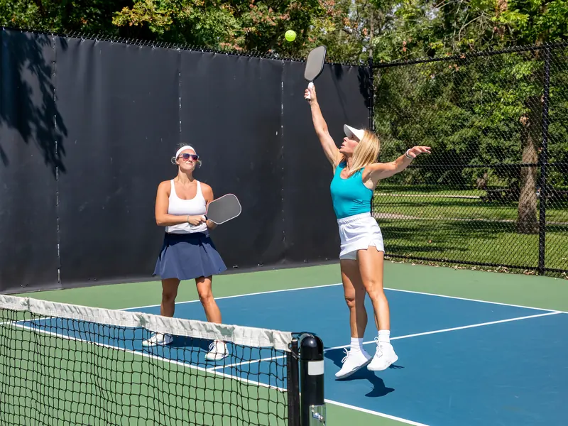Two women playing doubles pickleball, one leaping to serve as her partner watches