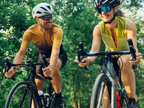 A smiling man and woman riding road bikes side by side through a leafy park