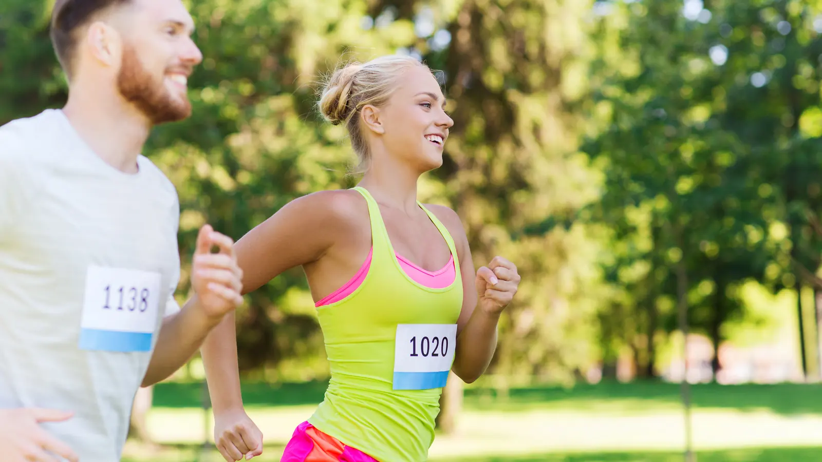 A smiling man and woman wearing race bibs running side by side through a sunny park