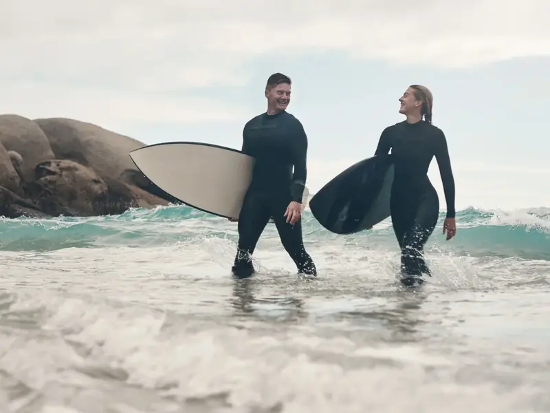 A smiling couple in wetsuits wading out into the surf carrying their surfboards