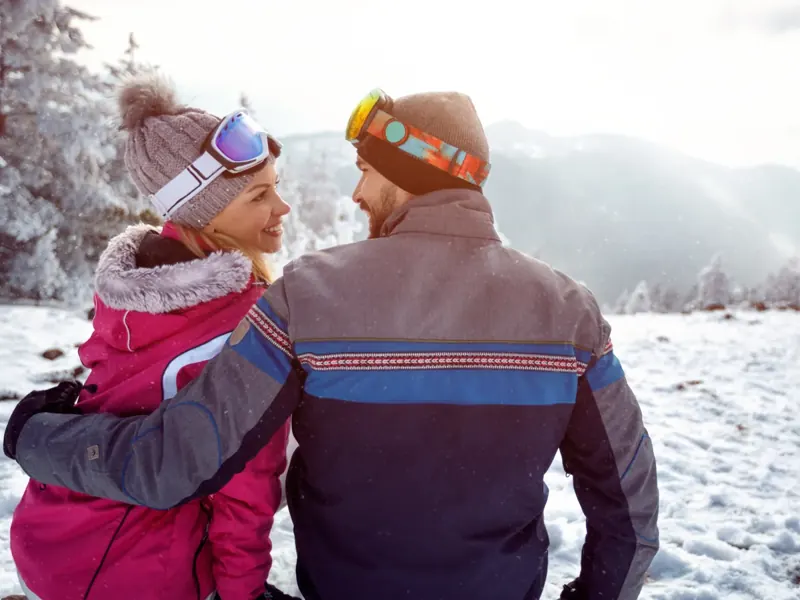 A couple in knit hats and goggles, arm in arm, smiling at each other in a snowy mountain landscape