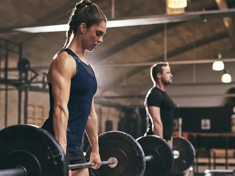 A woman and man setting up for barbell deadlifts in a warm, sunlit gym