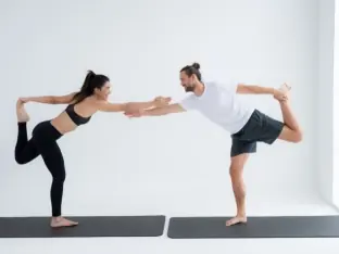 A man and woman holding hands in a partner balance yoga pose in a white studio