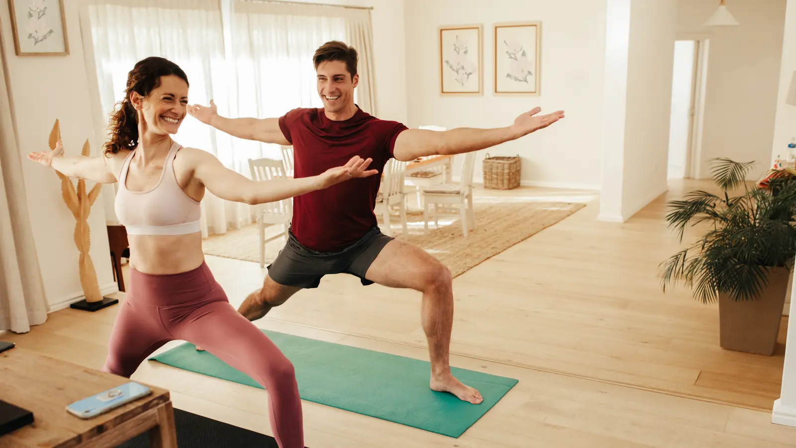 A smiling couple holding warrior pose side by side on yoga mats in a bright living room
