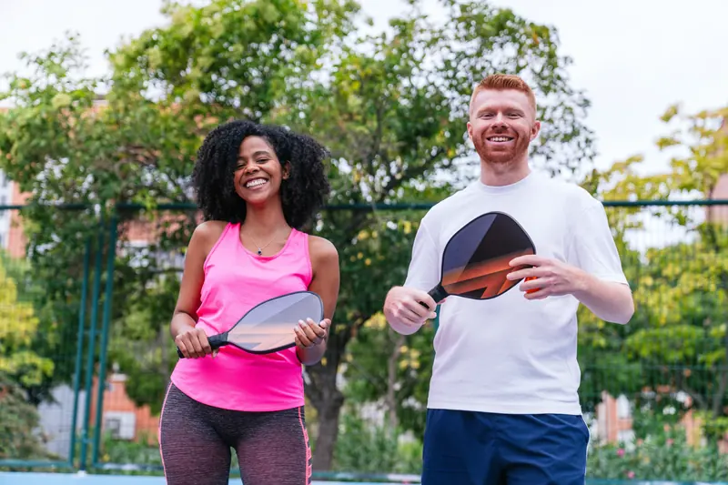 A smiling woman and man holding pickleball paddles on a sunny court