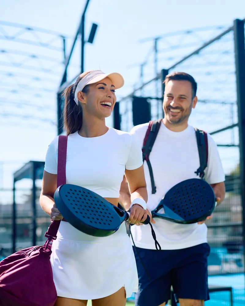 A laughing woman and man carrying padel rackets as they walk off a sunny court together