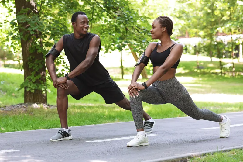 A smiling man and woman doing lunge stretches side by side on a sunny park path