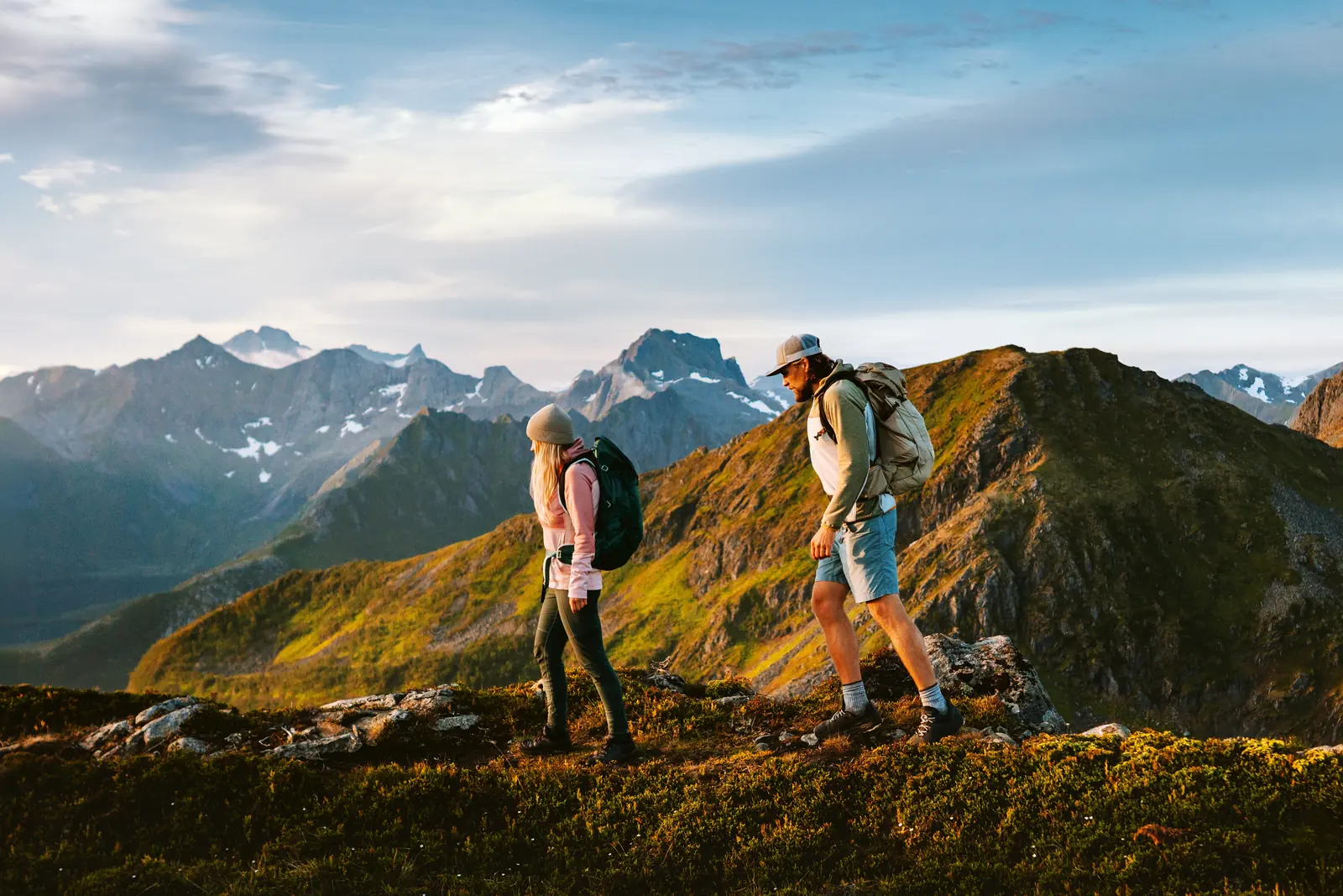 Two hikers walking along a sunlit mountain ridge with peaks behind them